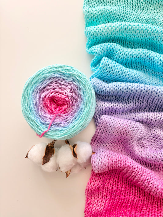 A knitted fabric swatch and matching yarn ball with a pastel gradient transitioning from turquoise to pink, shown on a light background with cotton bolls as styling props.